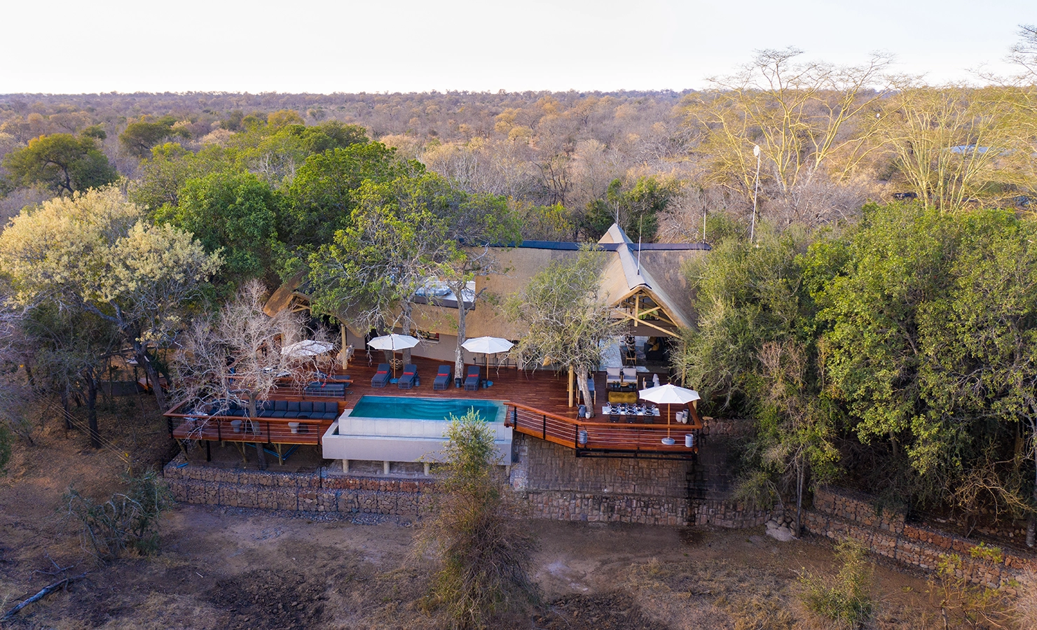 Perfect Hideaways: An aerial view of a luxury safari lodge nestled among lush trees, featuring a thatched-roof main building, expansive wooden deck, and an inviting infinity pool overlooking the bushveld. The lodge blends seamlessly into its natural surroundings, offering a tranquil retreat in the wilderness. Serondella, Thornybush Game Reserve.