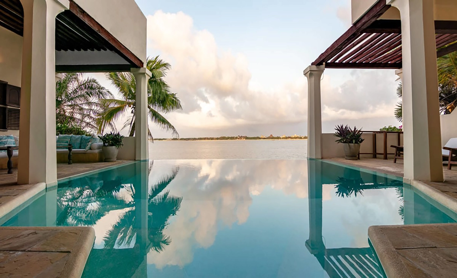 erfect Hideaways: A tranquil infinity pool reflecting the clouds, bordered by a shaded seating area and palm trees, overlooking the ocean. Shela Beach House, Lamu.