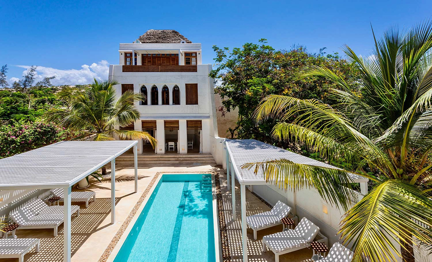 Perfect Hideaways: A vibrant daytime scene showcasing a villa surrounded by tropical greenery, with a central pool bordered by shaded loungers and lush palms. The Kasbah, Lamu.