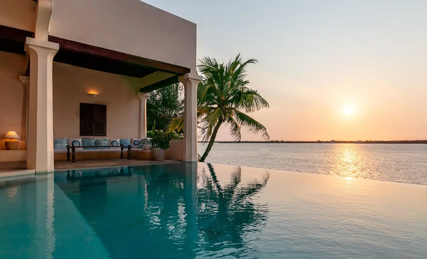 Perfect Hideaways: An infinity pool overlooking a serene ocean sunset, with a palm tree swaying beside a shaded lounging area. Shela Beach House, Lamu.