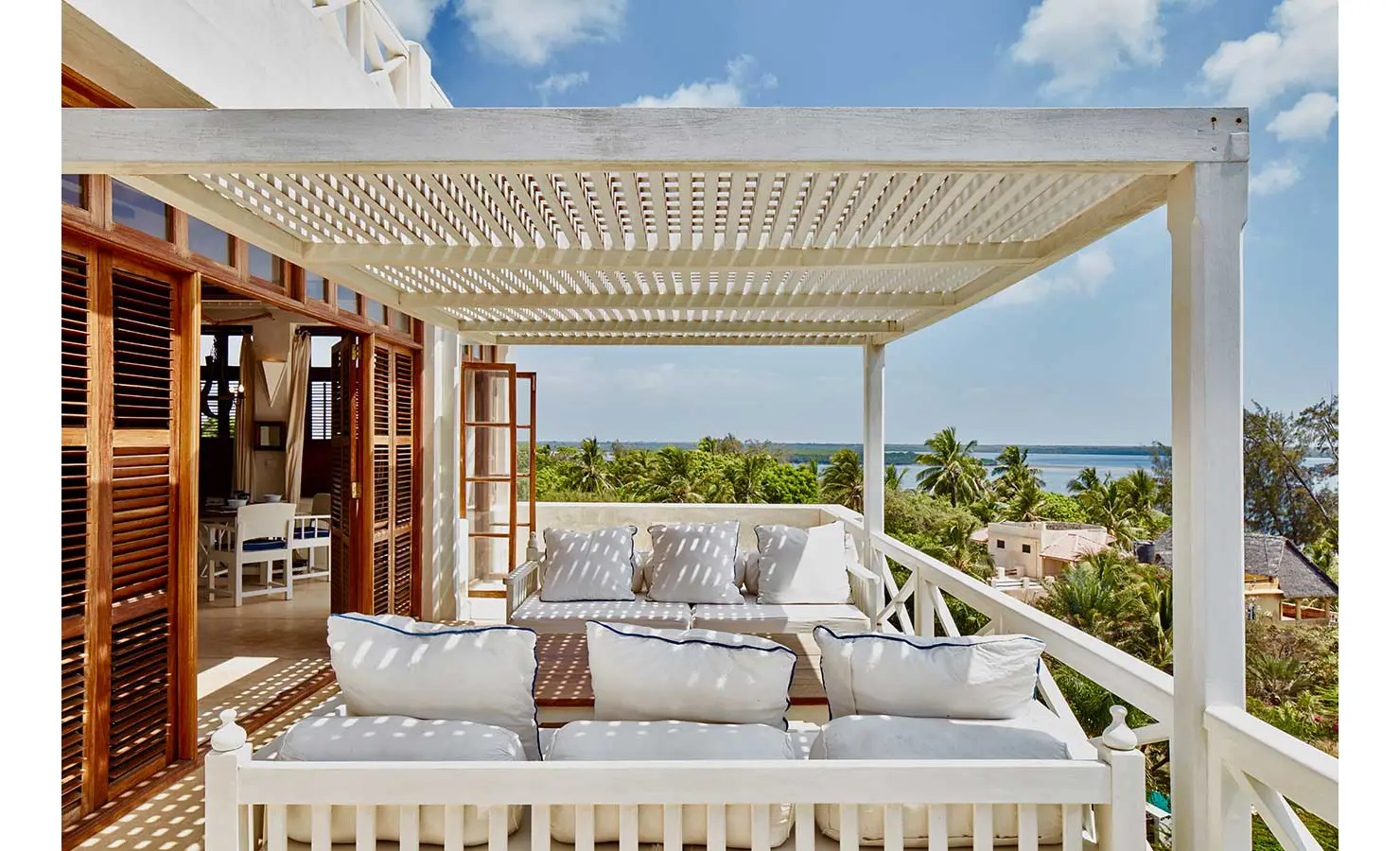Perfect Hideaways: A white pergola-covered terrace with plush white seating, overlooking lush palm trees and a distant view of the ocean. The Kasbah, Lamu.