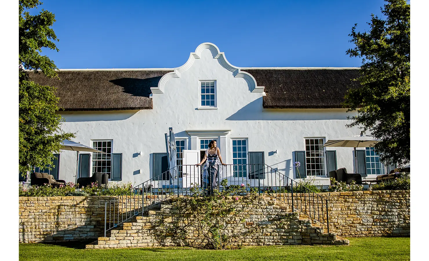 A lady standing on a split stone staircase landing with Brookdale, a whitewashed Cape Dutch manor house as the backdrop.
