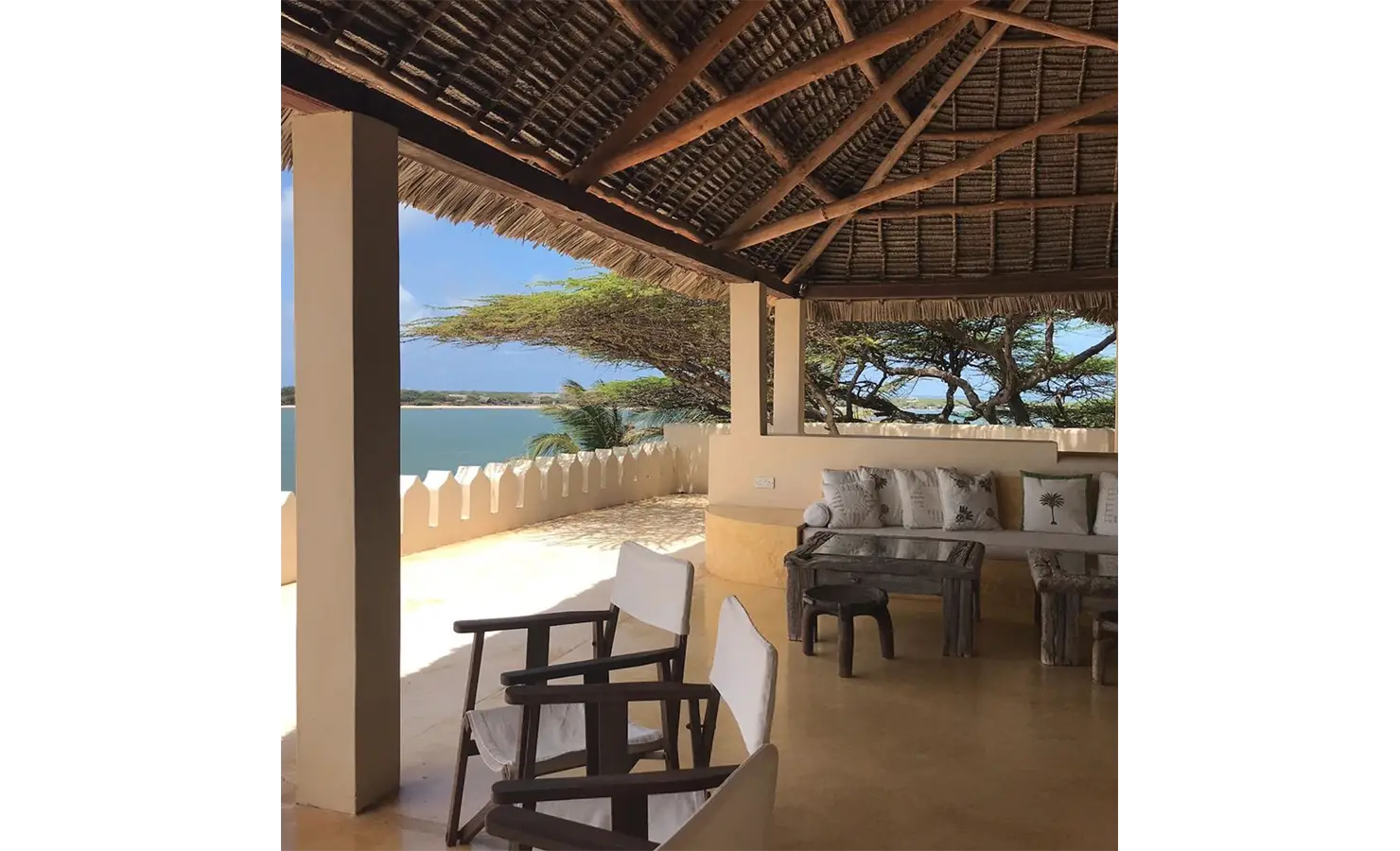 Perfect Hideaways: A breezy terrace with a thatched roof, cosy seating, and stunning ocean views framed by swaying trees. Shela Beach House, Lamu.