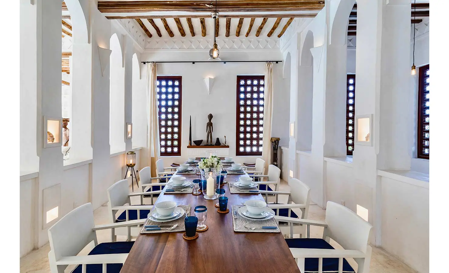 Perfect Hideaways: A beautifully set dining area with a long wooden table, white chairs, and intricate decorative niches, framed by arched windows and wooden beams. The Kasbah, Lamu.