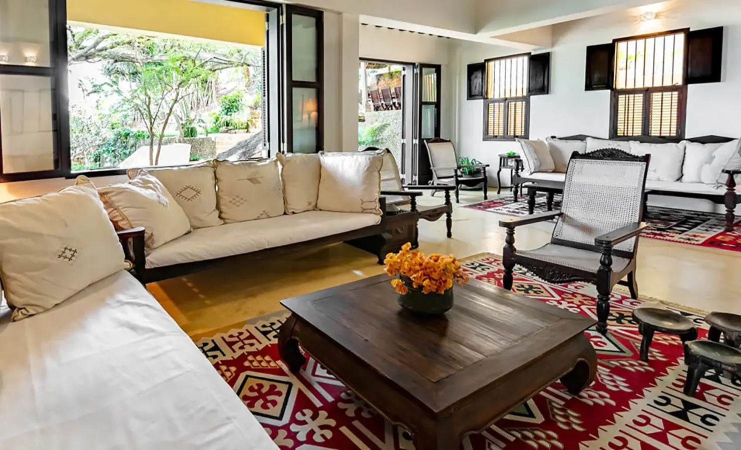 Perfect Hideaways: A bright and inviting living room with traditional Swahili furniture, vibrant patterned rugs, and open windows showcasing lush greenery. Shela Beach House, Lamu.