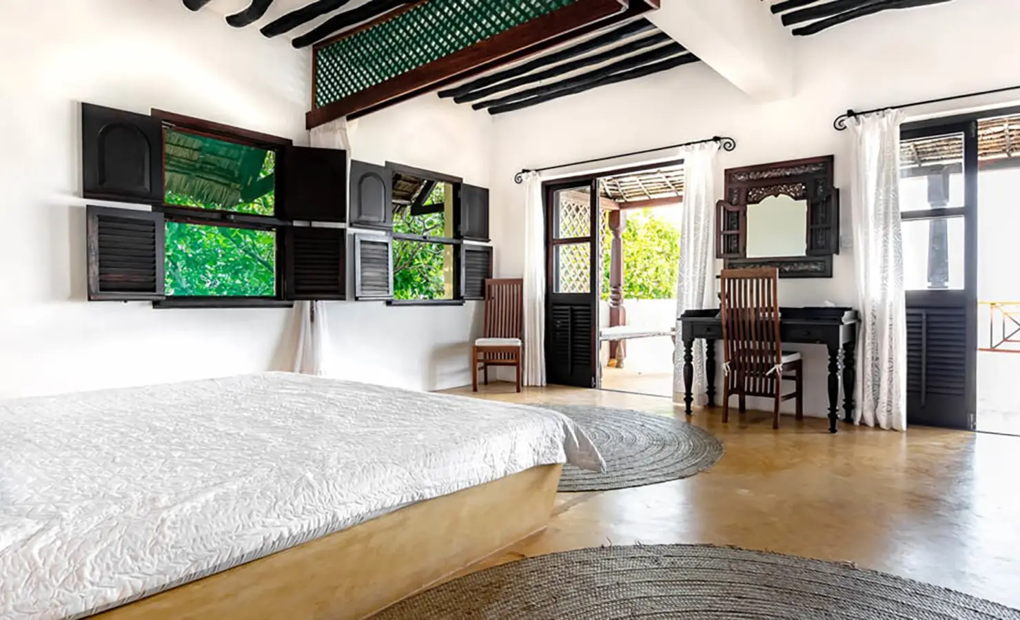 Perfect Hideaways: A spacious bedroom featuring white bedding, open wooden shutters revealing greenery, and traditional Swahili architectural details. Shela Beach House, Lamu.