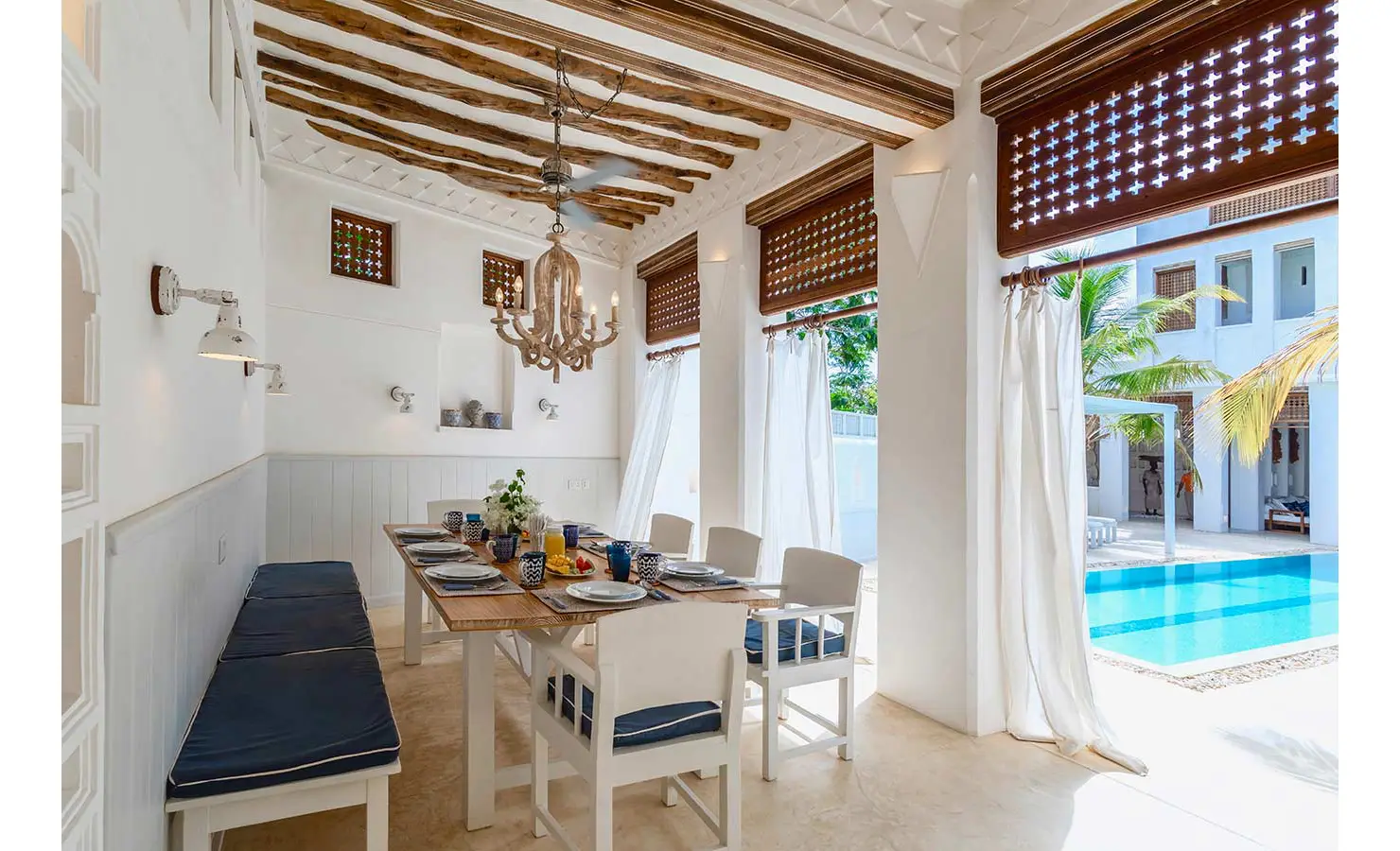 Perfect Hideaways: An open-air dining space with a rustic wooden ceiling, a chandelier, and a view of the pool surrounded by tropical greenery. The Kasbah, Lamu.