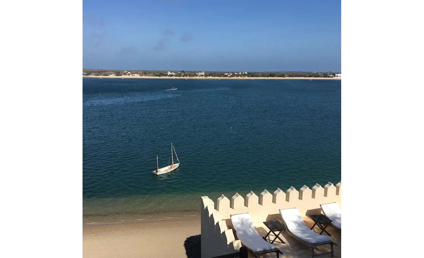 Perfect Hideaways: A breathtaking view of the ocean with white sandy shores, sun loungers, and a traditional dhow boat in the foreground. Shela Beach House, Lamu.