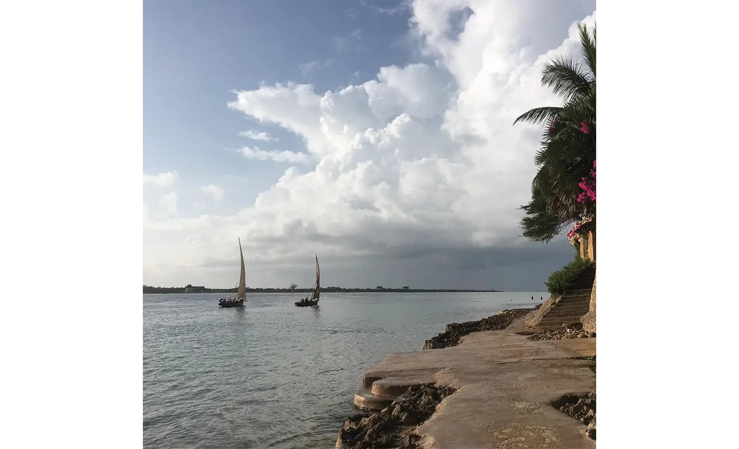 Perfect Hideaways: A tranquil scene of dhows sailing on calm waters under a dramatic cloudscape, bordered by lush greenery and floral accents. Shela Beach House, Lamu.