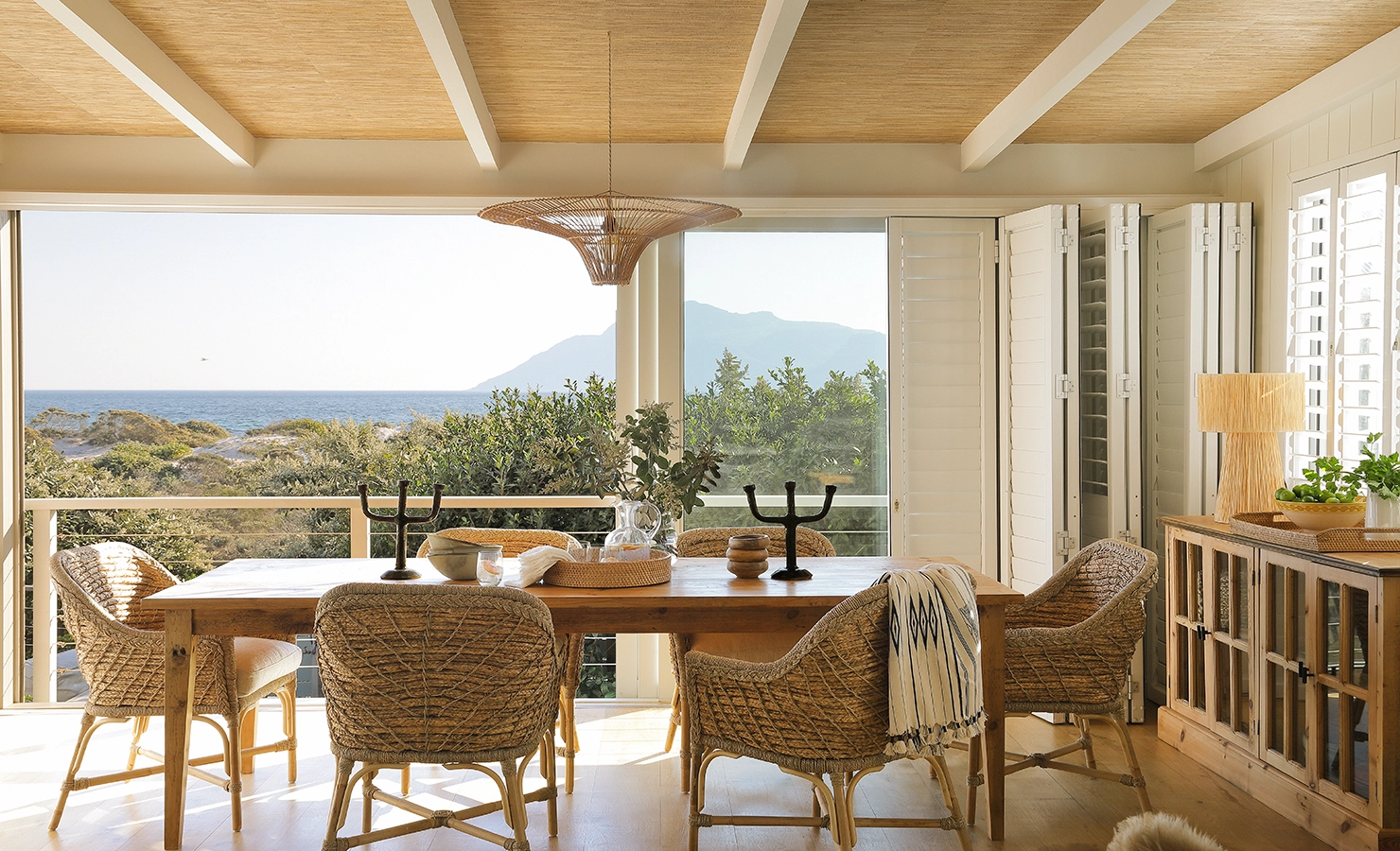 Perfect Hideaways: A serene dining space with woven rattan chairs and a rustic wooden table set against an open backdrop of ocean views and distant mountains. Natural textures, soft neutral tones, and an abundance of natural light create an inviting coastal retreat. Sunset Cove, Kommetjie.