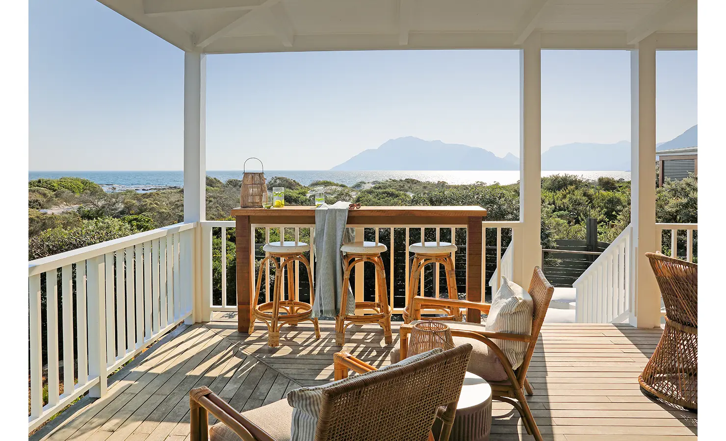 Perfect Hideaways: A breezy outdoor deck with a wooden bar table and rattan stools, framed by white railings and lush coastal greenery. The perfect spot for sipping sundowners while gazing at the sea. Sunset Cove, Kommetjie.