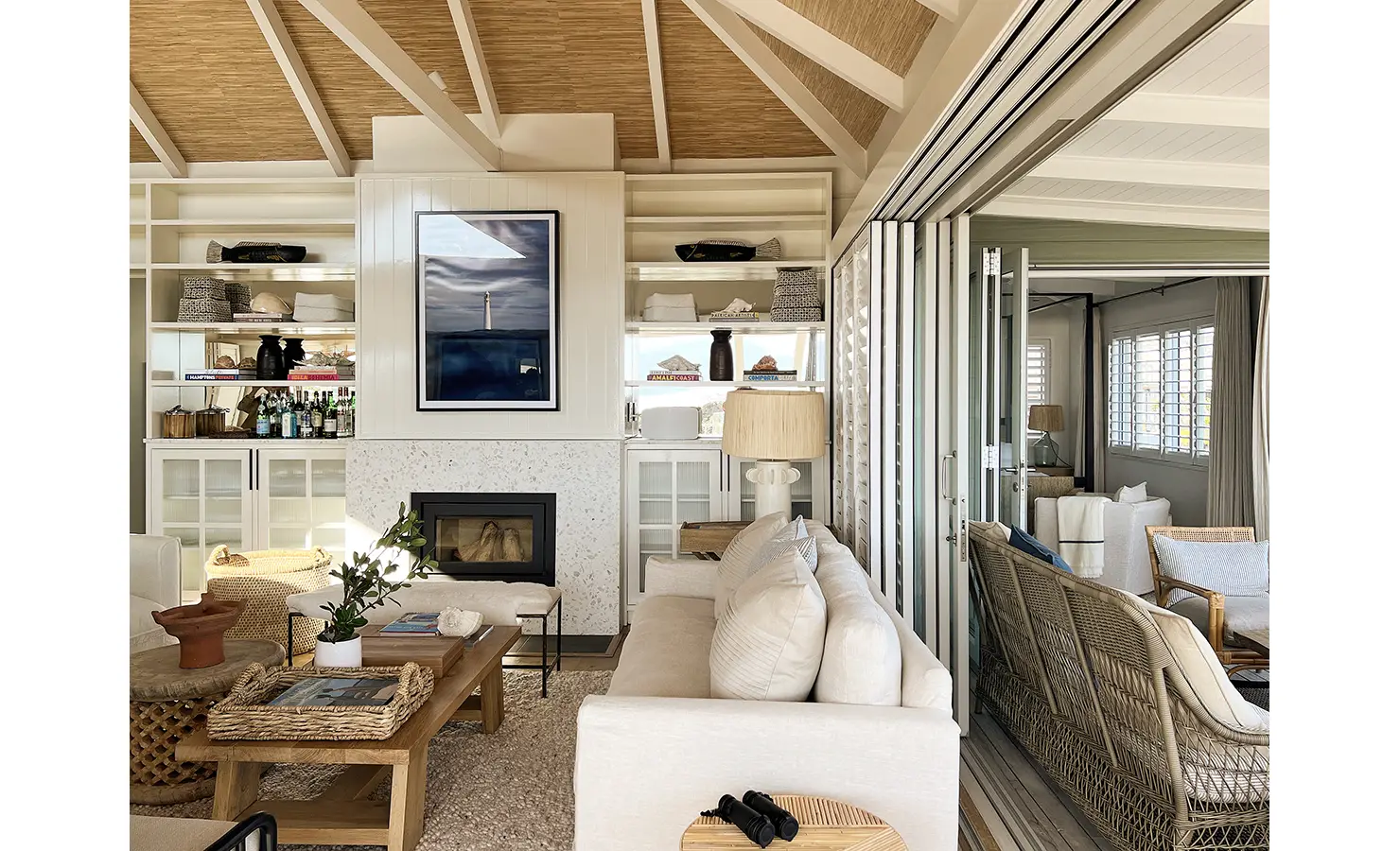 Perfect Hideaways: A chic, light-filled living area with a fireplace, woven textures, and a neutral palette. Bi-fold doors open onto a breezy deck, blurring the line between inside and out. Sunset Cove, Kommetjie.