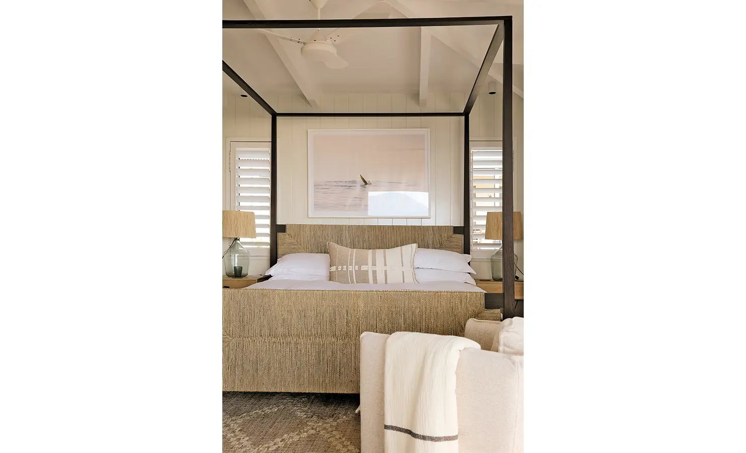Perfect Hideaways: A beautifully designed four-poster bed with woven textures and soft linen, framed by coastal artwork and warm lighting. A restful escape with understated elegance. Sunset Cove, Kommetjie.