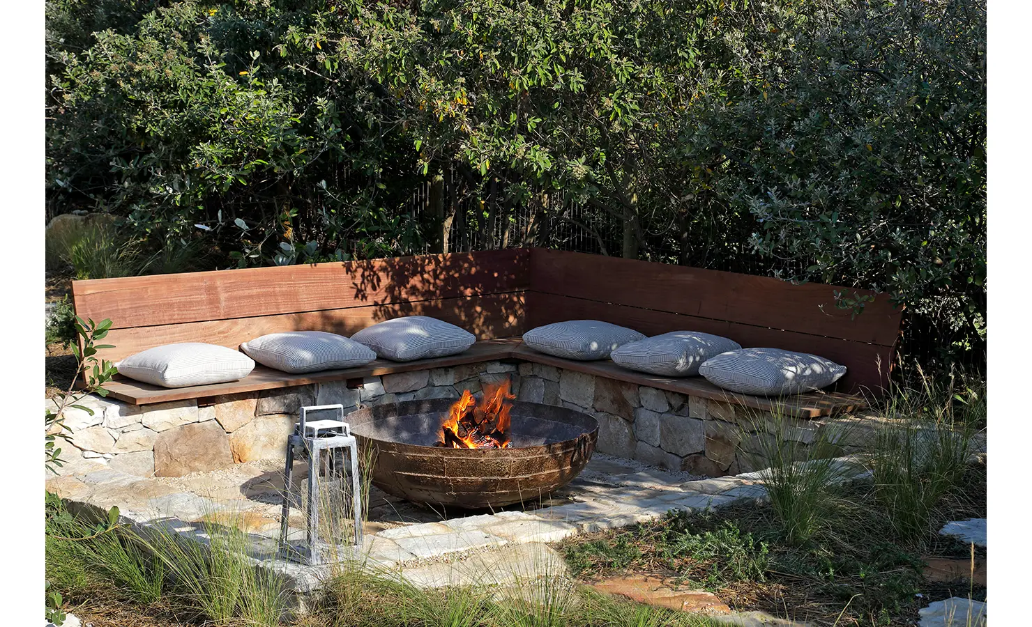 Perfect Hideaways: A secluded fire pit encircled by a stone and wood bench with soft cushions, nestled among lush greenery—a cosy retreat for outdoor gatherings under the stars. Sunset Cove, Kommetjie.