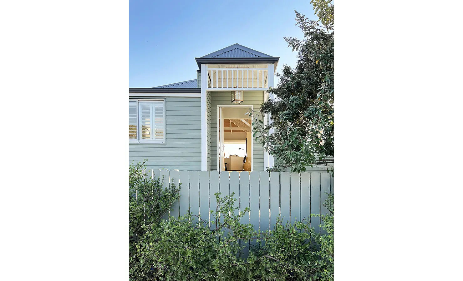 Perfect Hideaways: A charming coastal home with sage green cladding and white trim, framed by lush foliage. The inviting entrance leads into an airy interior with ocean views beyond. Sunset Cove, Kommetjie.