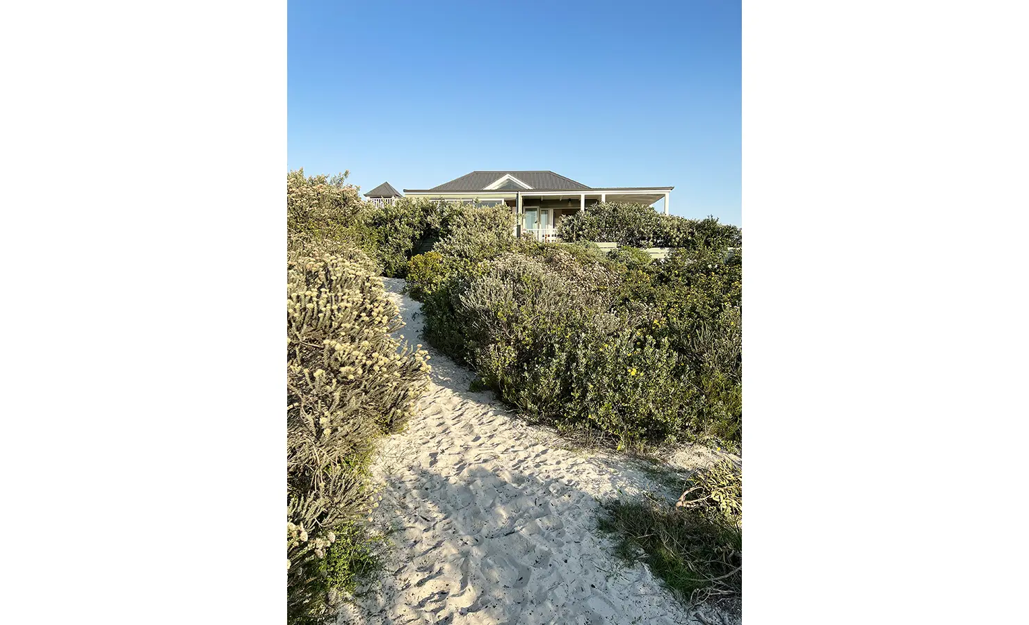 Perfect Hideaways: A sandy footpath winds through native coastal vegetation, leading to a serene beach house tucked among the dunes—a true hideaway by the sea. Sunset Cove, Kommetjie.