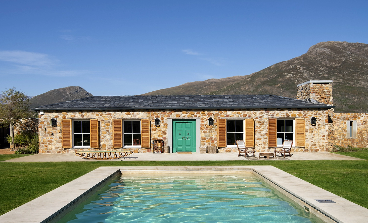Perfect Hideaways: A charming stone-clad cottage with wooden shutters and a striking turquoise door, set against the backdrop of rolling mountains. A sparkling swimming pool in the foreground reflects the clear blue sky, while outdoor seating invites relaxation on the terrace. Drakenskloof, Wellington.