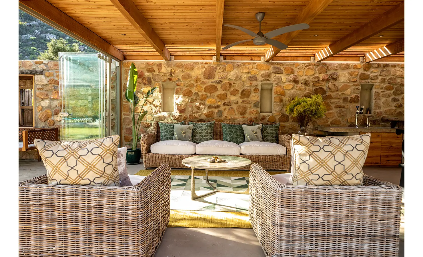 Perfect Hideaways: A stylish open-plan lounge with wicker armchairs, a rattan sofa, and green accent pillows, set against a rustic stone wall. Sunlight filters through the slatted wooden ceiling, enhancing the inviting atmosphere. Drakenskloof, Wellington.