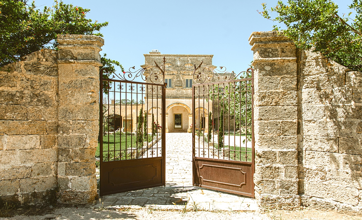 Perfect Hideaways: A grand entrance framed by ornate wrought-iron gates, revealing a stone pathway leading to an elegant historic villa surrounded by lush gardens. Villa Tafuri, Nardo.