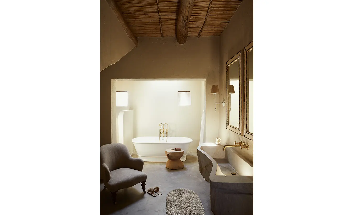 Perfect Hideaways: A warm-toned bathroom with a freestanding white bathtub, a stone sink, and a cosy armchair under a thatched ceiling. Sterrekopje, Franschhoek.