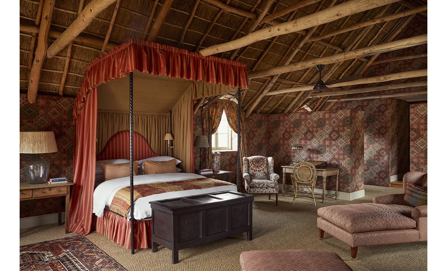 Perfect Hideaways: A lavish bedroom with a red-canopied bed, patterned wallpaper, and a rustic thatched ceiling, featuring plush seating and a carved wooden chest. Sterrekopje, Franschhoek.