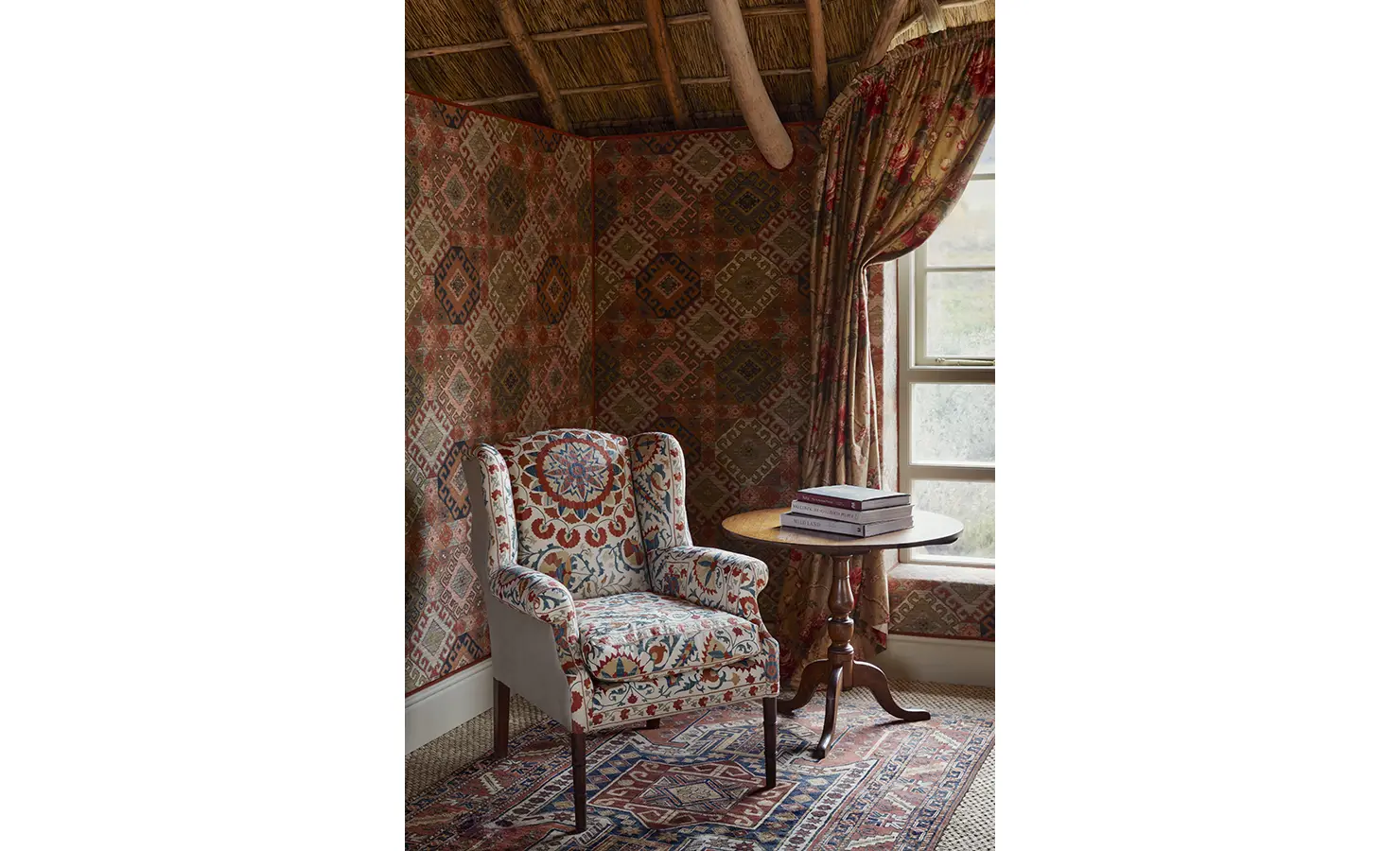 Perfect Hideaways: A cosy corner with a vibrant patterned armchair, a round wooden table, and floral drapes under a thatched ceiling. Sterrekopje, Franschhoek.