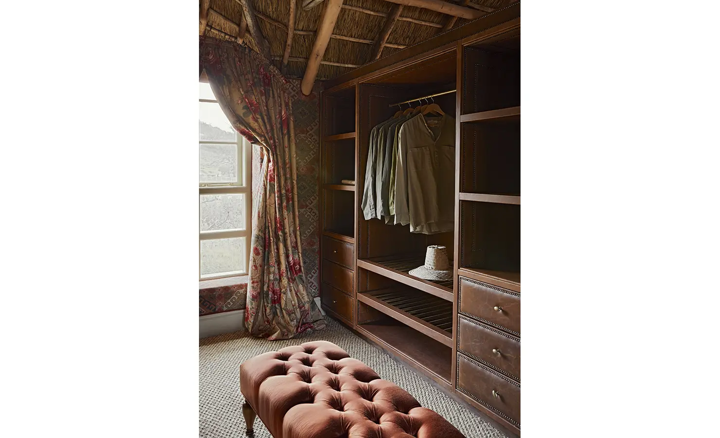 Perfect Hideaways: A walk-in wardrobe with dark wooden shelving, floral drapes, and a tufted ottoman under a thatched ceiling. Sterrekopje, Franschhoek.