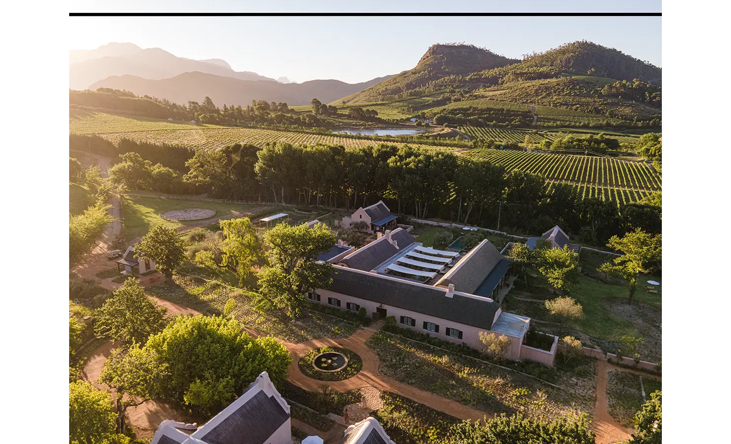 Perfect Hideaways: An aerial view of the estate surrounded by lush vineyards, rolling hills, and a central Cape Dutch-style farmhouse. Sterrekopje, Franschhoek.