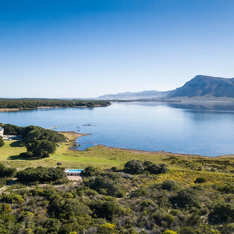 Perfect Hideaways: Panoramic aerial of the Klein River Lagoon bordered by fynbos and lawns, with a lagoon-side pool and distant mountain ridges beneath a clear blue sky, Stanford, Coot Club.
