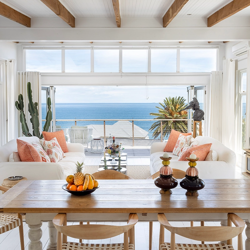 Perfect Hideaways light-filled open-plan living and dining area with white sofas, coral accent cushions, exposed wooden beams, and large glass doors opening onto a sea-facing balcony with palm trees and uninterrupted ocean views, Summer Rose, Camps Bay