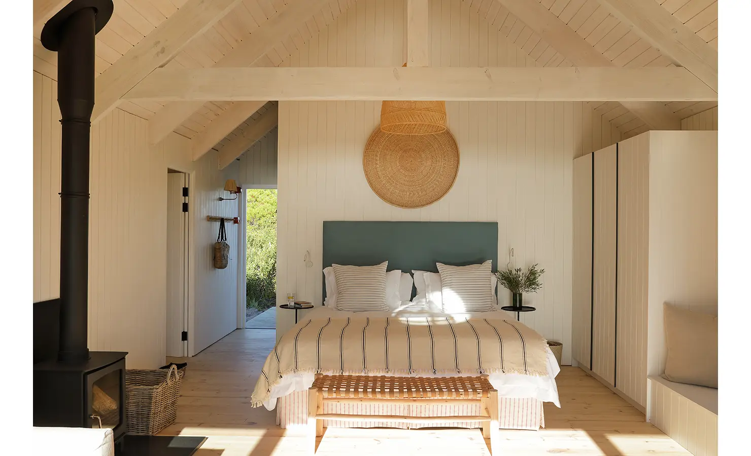Perfect Hideaways: A serene bedroom with vaulted ceilings, a wicker bench at the foot of the bed, and a woven wall accent above a teal headboard. Natural light fills the room. Coot Club, Stanford.