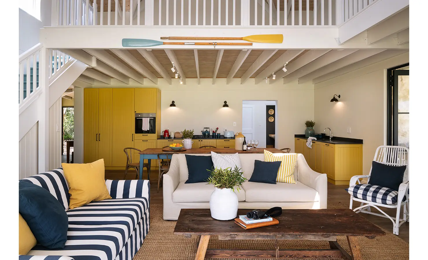 Perfect Hideaways: Open-plan living area with double-volume ceiling and mezzanine, bold yellow kitchen cabinetry, striped navy sofa and decorative oars, Stanford, Coot Club.