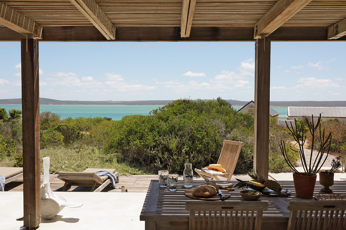 Perfect Hideaways: Slatted-roof terrace with an alfresco dining table set for lunch, overlooking sun loungers, fynbos and the turquoise sweep of Langebaan Lagoon under bright skies, Lagoon House, Churchhaven.