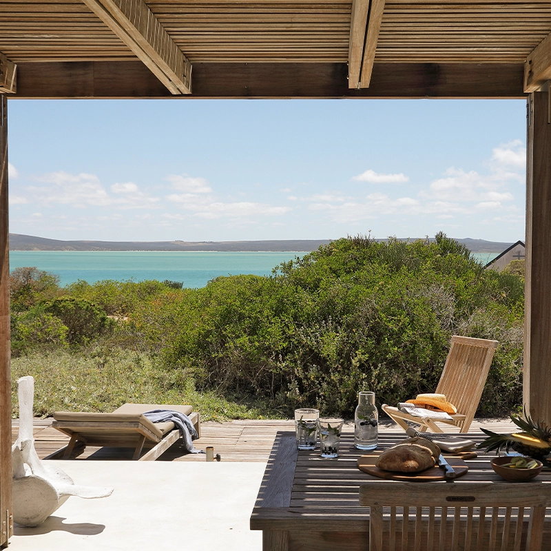 Perfect Hideaways: Slatted-roof terrace with an alfresco dining table set for lunch, overlooking sun loungers, fynbos and the turquoise sweep of Langebaan Lagoon under bright skies, Lagoon House, Churchhaven.