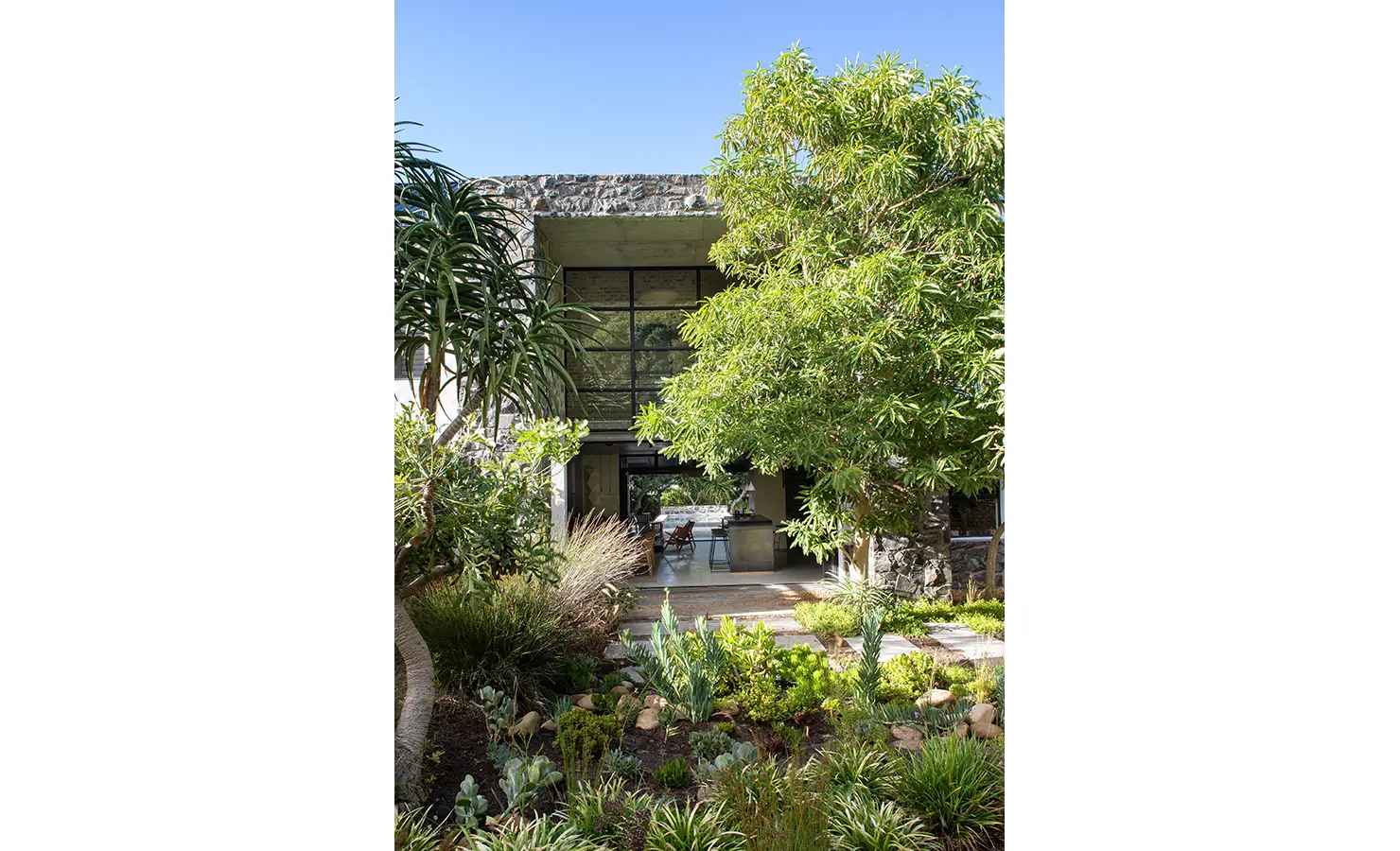 A landscaped garden with indigenous plants leading to a contemporary stone-clad house with floor-to-ceiling glass windows, blending seamlessly with nature. Pinebeach, Noordhoek.