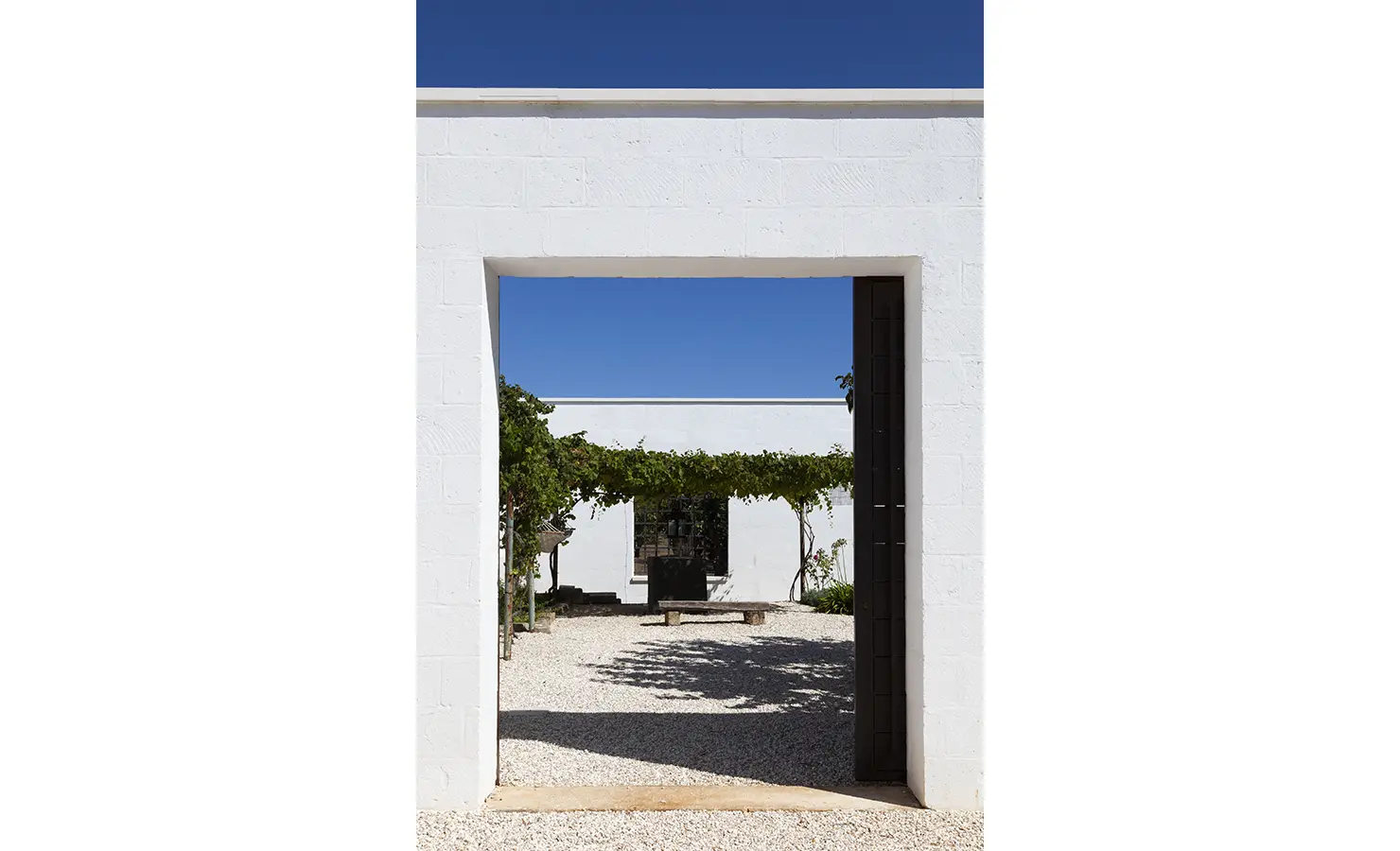 Perfect Hideaways: A sun-drenched courtyard framed by crisp white walls, featuring a wide entrance that opens to a tranquil outdoor space with a shaded pergola draped in vines. The deep blue sky contrasts beautifully with the minimalist architecture. La Casetta, Ostuni.