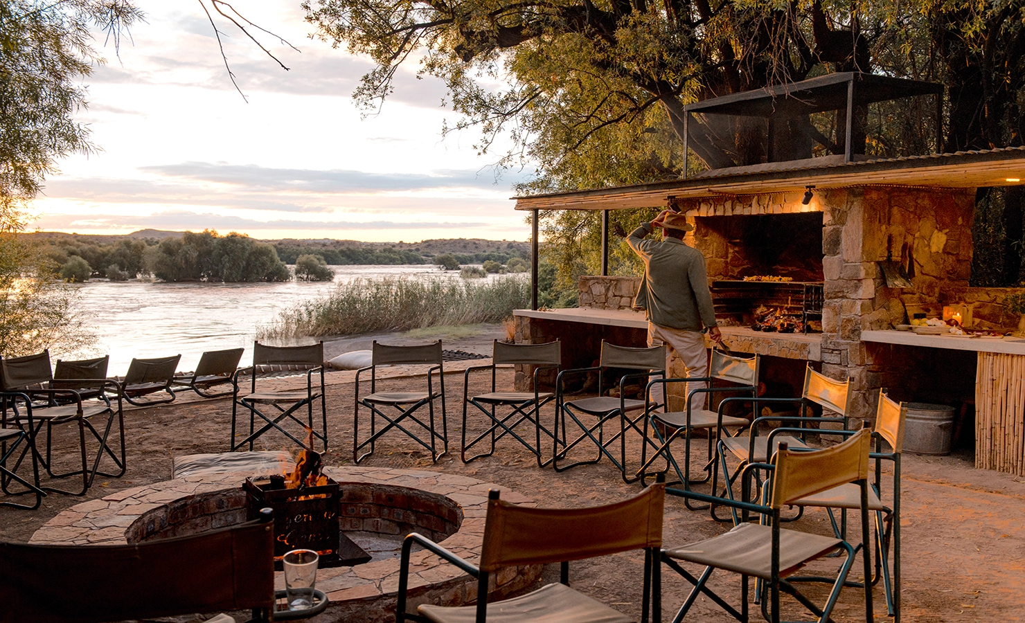 Perfect Hideaways: An idyllic riverside braai area at sunset, featuring a firepit surrounded by canvas chairs and a rustic outdoor kitchen with a stone-built fireplace. A man in a hat tends to the grill, as the Orange River flows peacefully in the background, framed by lush greenery and distant hills. Orange River House, Hopetown.
