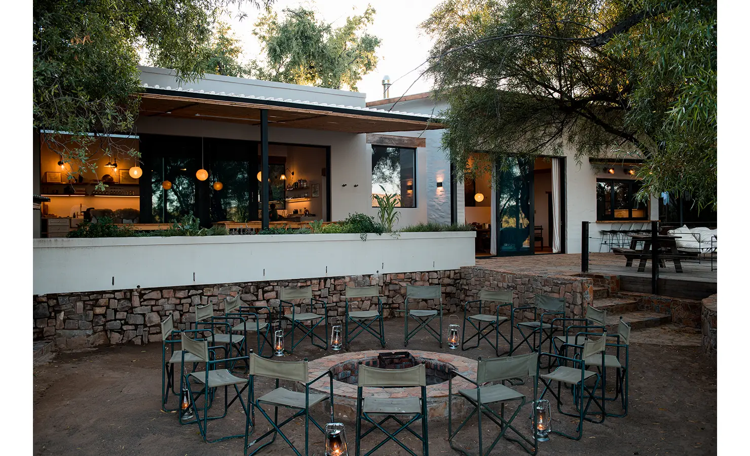 Perfect Hideaways: A firepit encircled by canvas chairs sits at the heart of an inviting outdoor gathering space, with glowing lanterns adding warmth as evening falls. Behind, a modern home with floor-to-ceiling glass windows radiates a cosy ambiance, seamlessly blending with its natural surroundings. Orange River House, Hopetown.