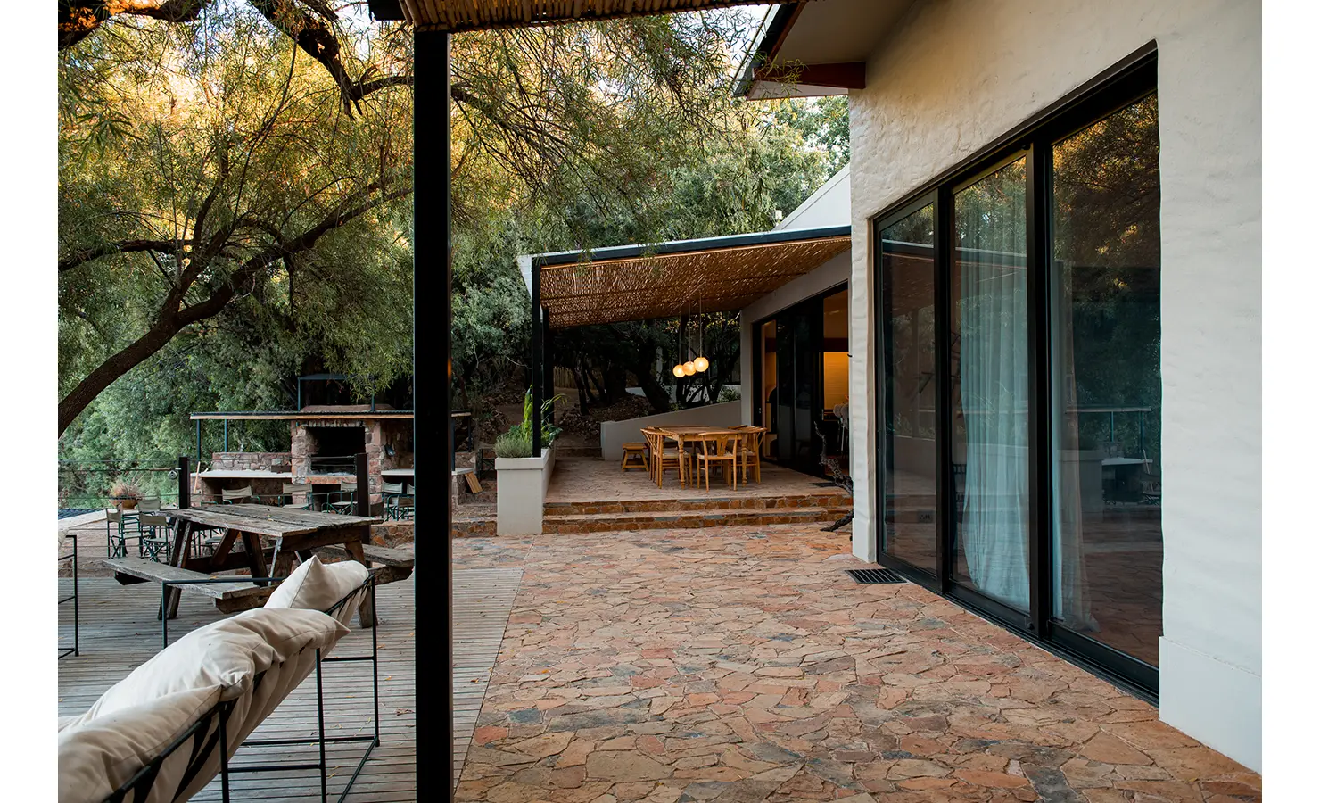 Perfect Hideaways: A serene outdoor patio with stone paving leads to a shaded dining area under a wooden pergola, offering a seamless indoor-outdoor flow. A rustic braai area sits nearby, framed by lush trees, overlooking the tranquil river beyond. Orange River House, Hopetown.