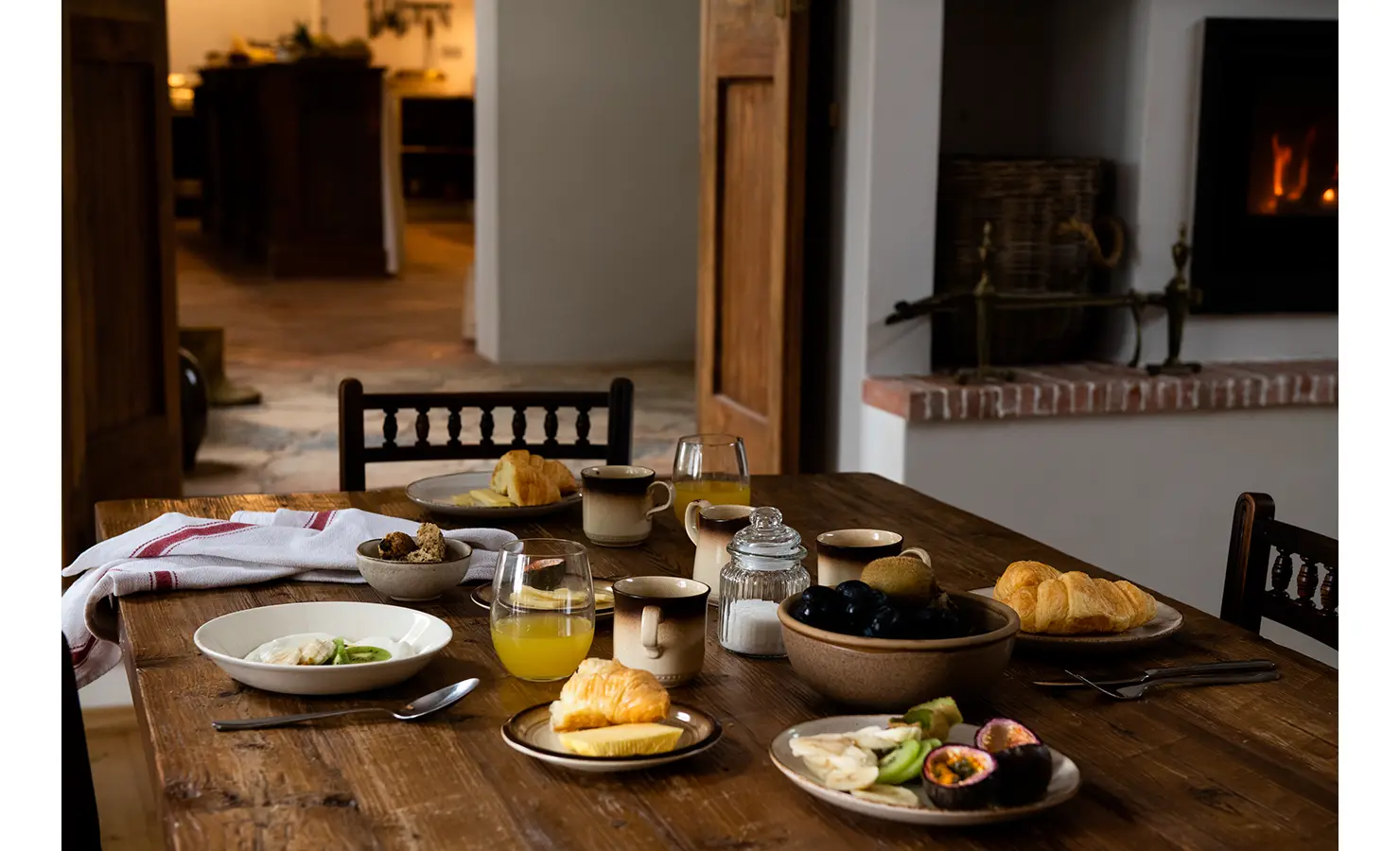 Perfect Hideaways: A beautifully set wooden dining table is adorned with fresh fruit, croissants, and steaming coffee, inviting a leisurely breakfast by the warm glow of a fireplace in the background. Orange River House, Hopetown.