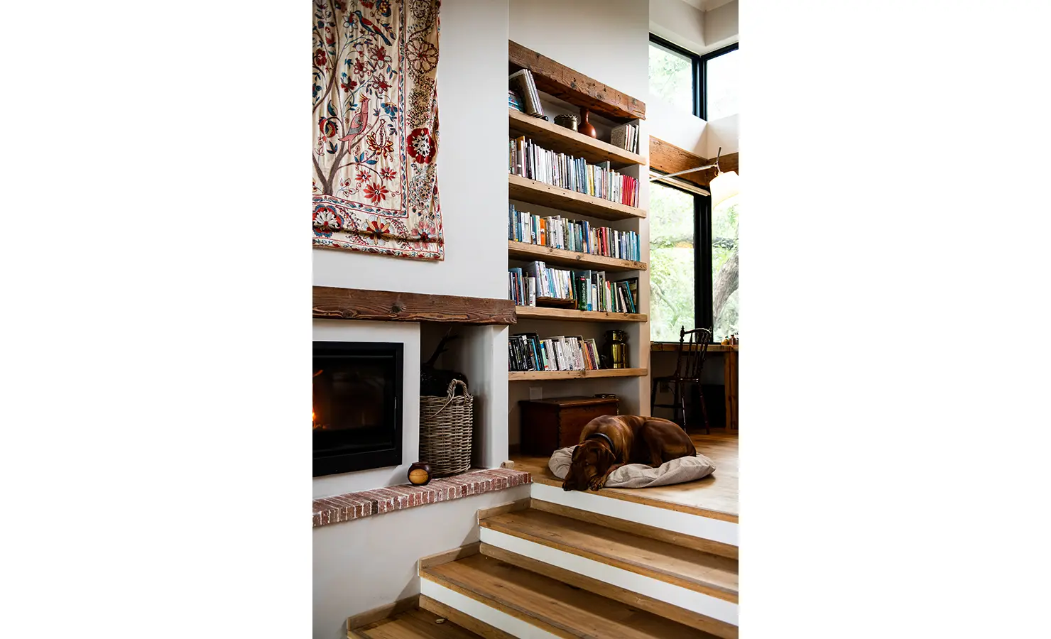 Perfect Hideaways: A cosy reading nook with a built-in wooden bookshelf, a roaring fireplace, and a sleeping dog creates a serene retreat. Sunlight streams through large windows, enhancing the inviting atmosphere. Orange River House, Hopetown.
