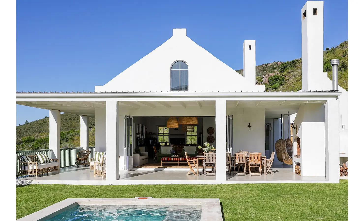 Buchubos Villa wrap around verandah with an outdoor wooden dining table, wicker sofa's and hanging wicker chairs with an outdoor braai, and a white finish.