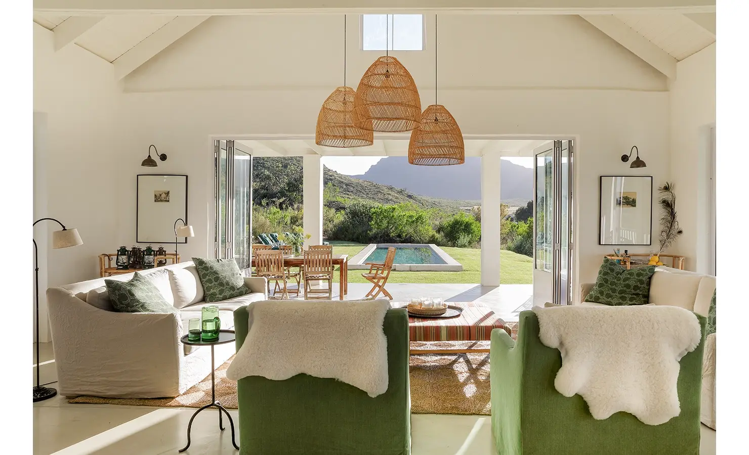 Open plan living area with a soft, white linen sofa, central hanging woven lights and two green armchairs that compliments the green lawn and surrounding landscape that is exposed via large glass stack back doors. Buchubos Villa.