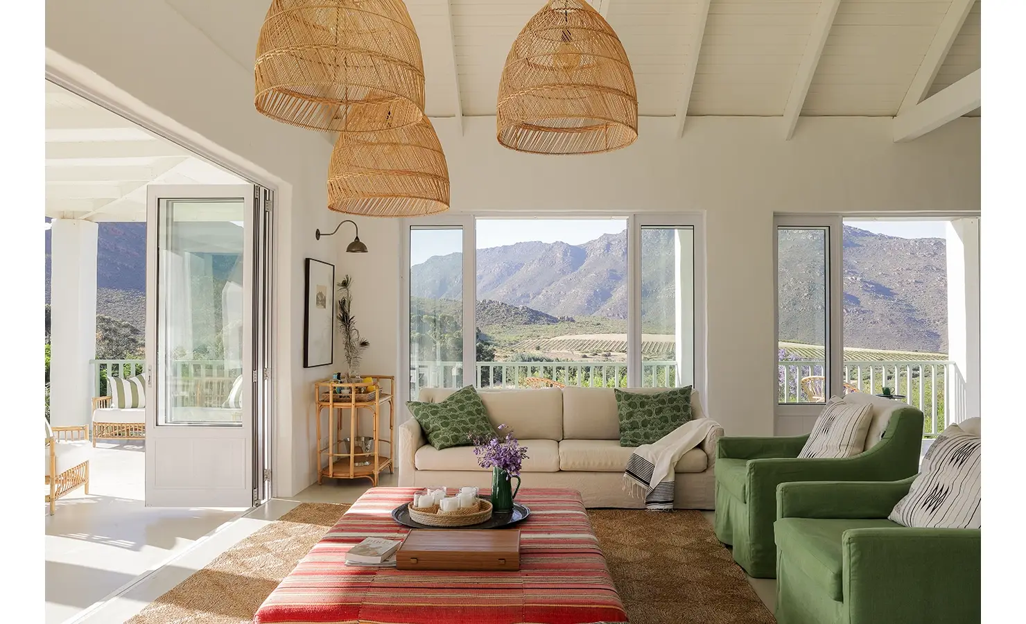 Buchubos living area with a soft beige sofa, green armchairs, a striped ottoman positioned below woven hanging lights and surrounded by large windows and stack doors exposing the mountain range.