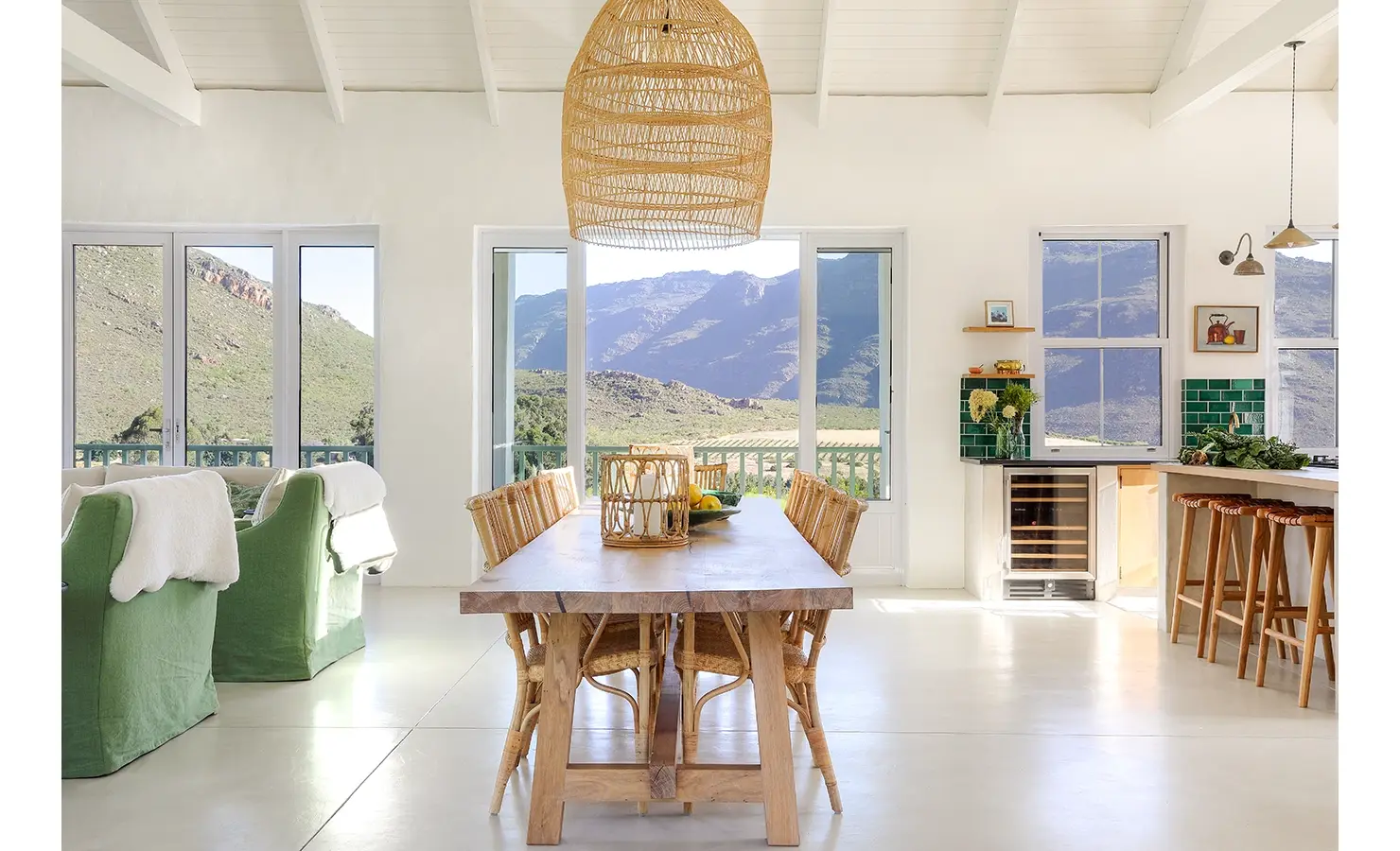 Open plan lounge and dining area with long wooden dining table and wicker chairs facing large glass windows with expansive views of the mountain range.