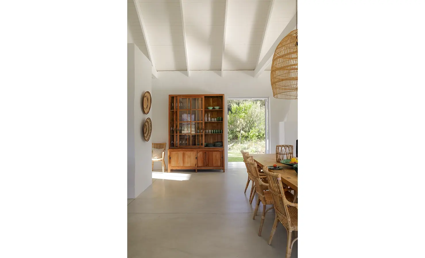 Open plan dining space with beige epoxy flooring and white walls, and a wooden dining table that compliments the large wooden framed glass cabinet. Buchubos Villa.