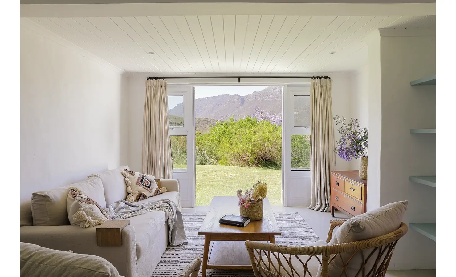 Secluded lounge with a soft, beige sofa, a wooden coffee table and wooden cabinet with double doors that open onto the luscious green garden of Buchubos Villa.