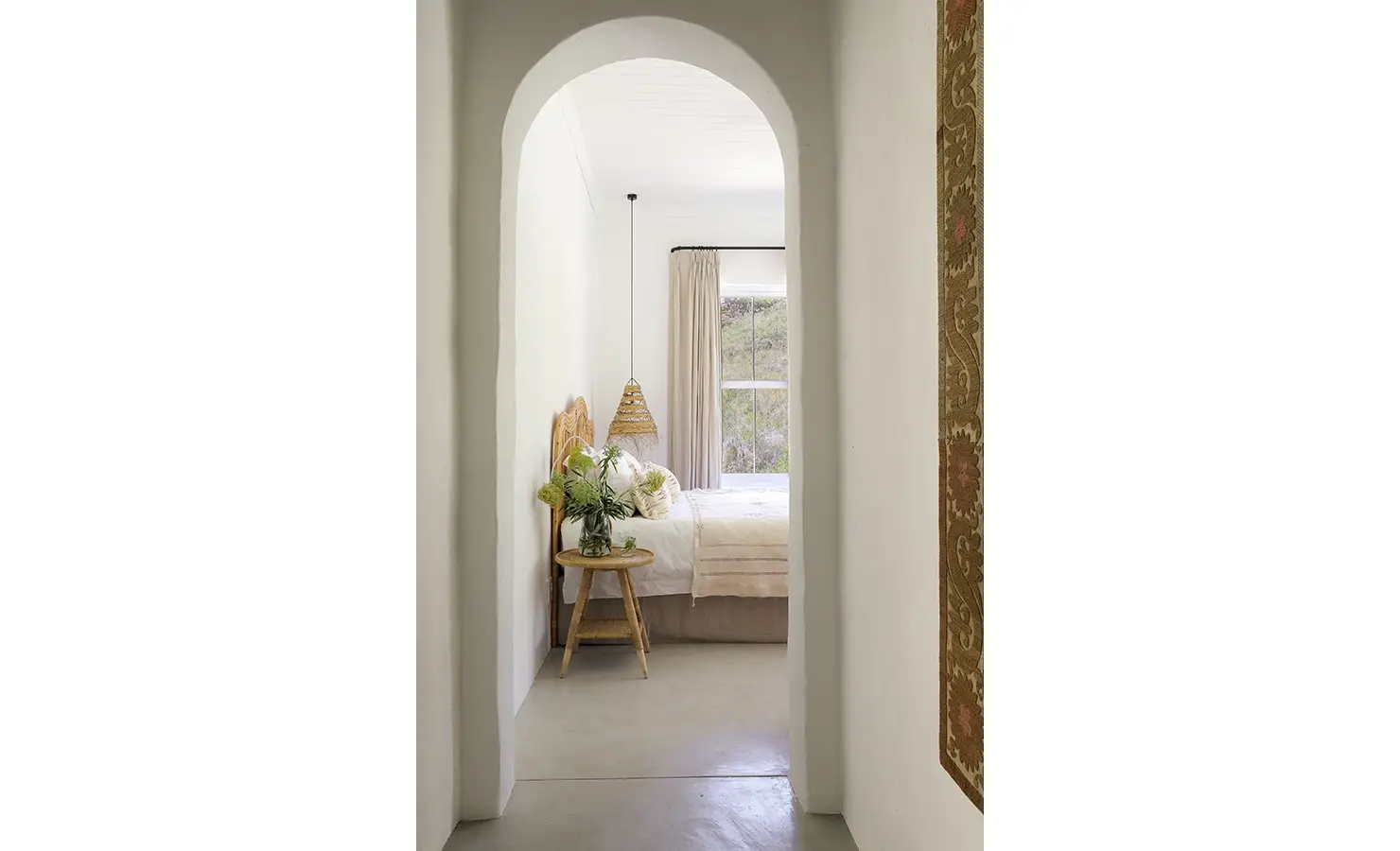 Arched entrance with whitewashed walls revealing a spacious bed with white linen, a wicker headboard and side table. Buchubos Villa.