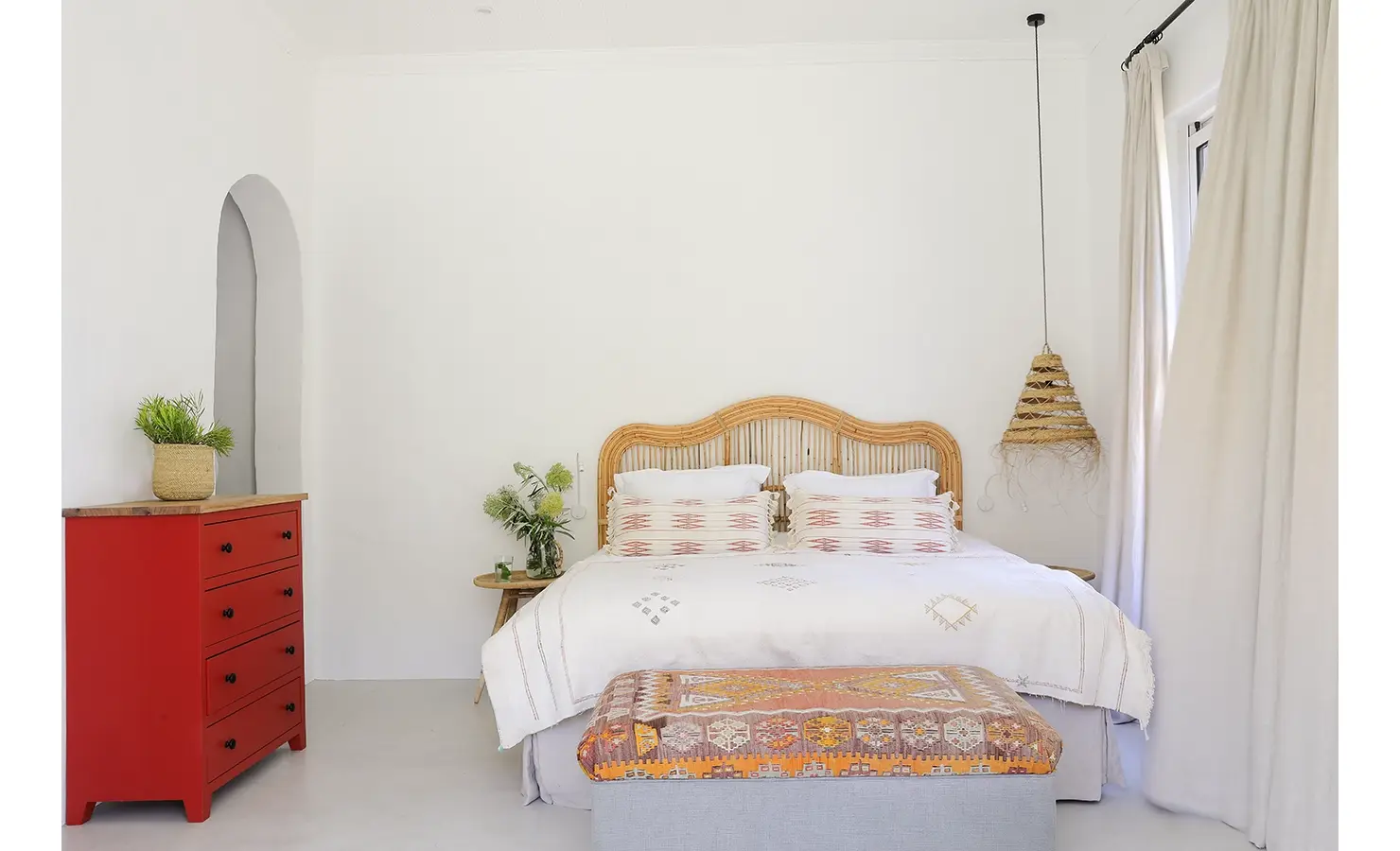 Buchubos Villa bedroom with whitewashed walls, a spacious bed with white linen and touches of pink attached to a wicker headboard, a side table and a colourful fabric ottoman at the head of the bed, with a red statement cabinet.