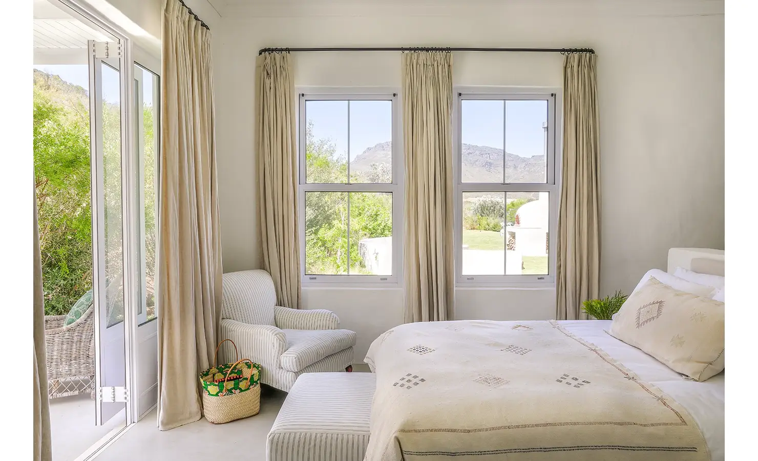 Comfortable bedroom with beige hues, a spacious bed with white linen and beige throw, a striped corner armchair and surrounding windows and double doors with views of Buchubos villa garden.